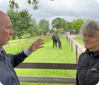 Iris and a man standing outdoors in a grassy area with a horse in the background